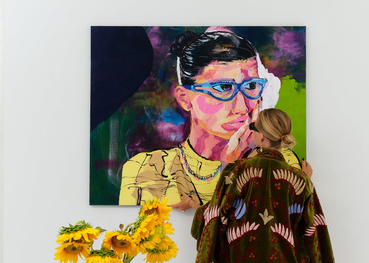 A person stands in a dining area wearing a dark green embroidered jacket with pink and blue details, facing a colorful portrait painting on a white wall. A light blue table with matching chairs and a vase of yellow sunflowers are in the foreground.